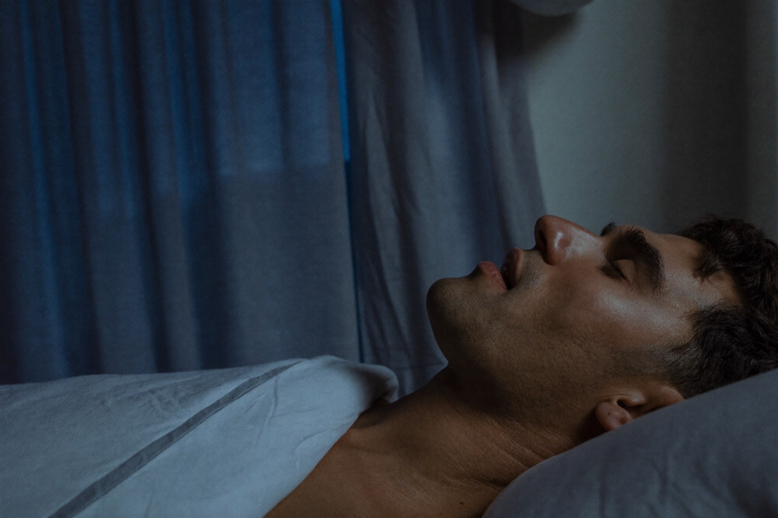 A man asleep on his back in dim light, head tilted back and mouth open.