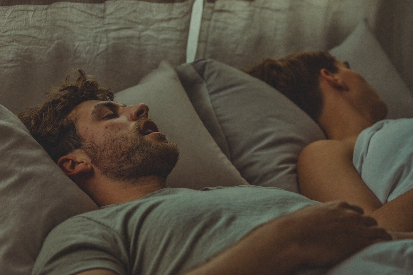 A man asleep on his back with his mouth open, his partner turned away beside him.