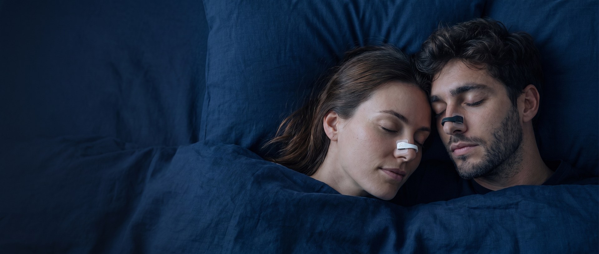 A couple asleep in bed, each wearing an Airlift magnetic nasal clip, black on him and white on her